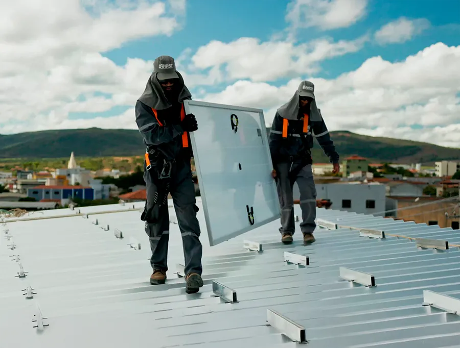 Dois instaladores da HASE caminhando sobre o telhado metálico carregando um painel solar, com os trilhos de fixação já montados.