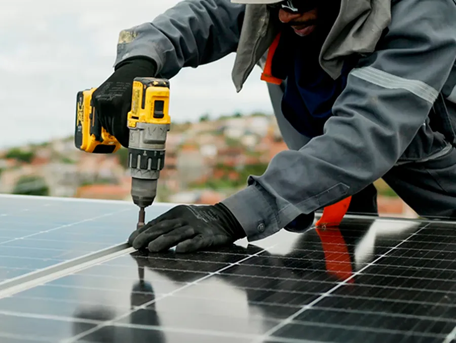 Mãos enluvadas de um técnico da HASE aparafusando com furadeira a fixação de um painel solar no trilho.