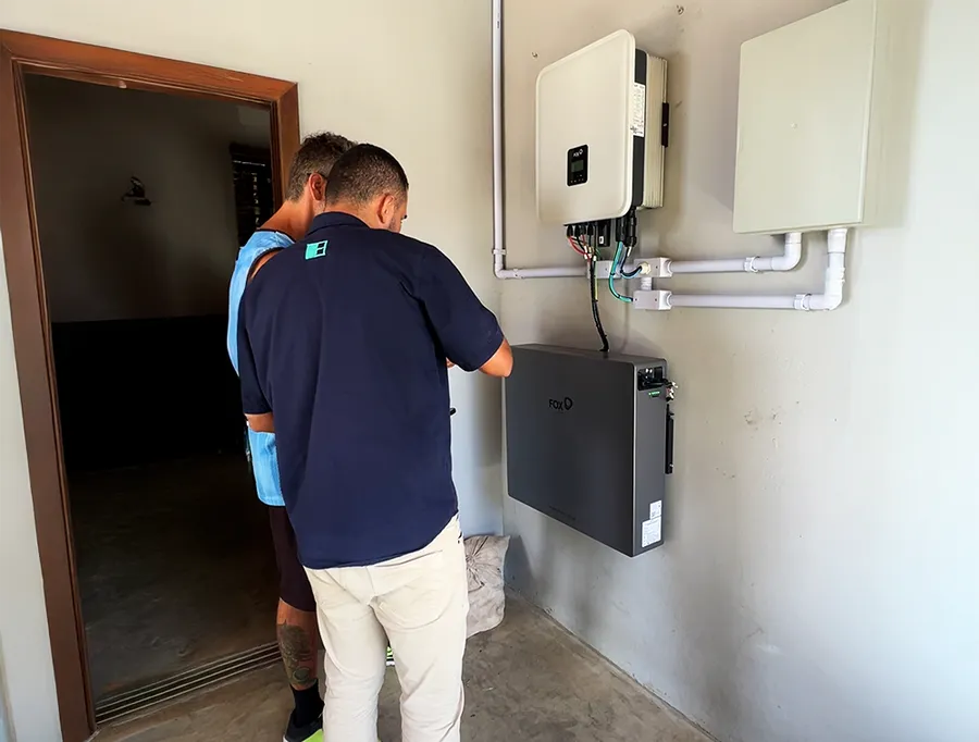 Técnico da HASE ao lado do cliente diante do inversor e da bateria recém-instalados na parede do imóvel.