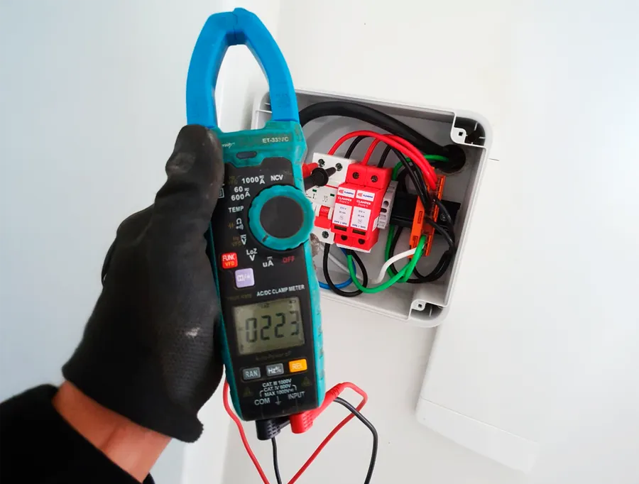 Técnico da HASE medindo a corrente com alicate amperímetro no quadro de proteção da usina, com disjuntores e DPS à mostra.